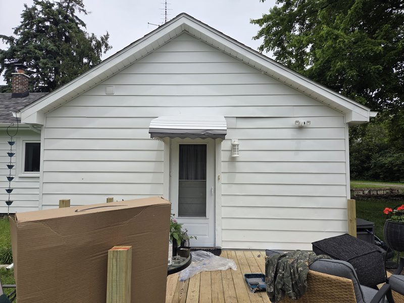 Exterior house painting, white siding and trim around a back door. A paint tray and plastic drop cloth sit on the deck.
