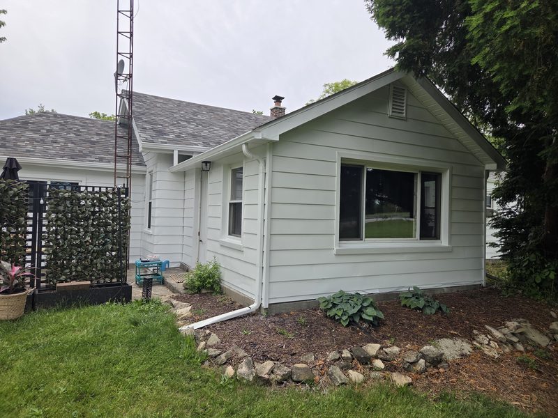 Exterior house painting, white siding and trim. Side of a one-story house with a large window in the gable end, white gutters and a stone-edged garden bed in front.
